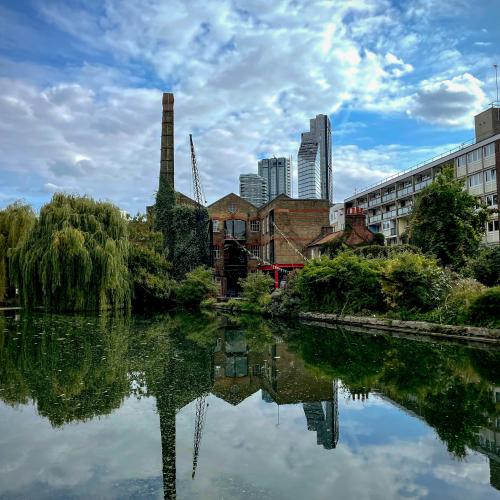 London canal reflecting willow trees, a brick warehouse, and distant skyscrapers.